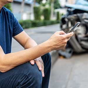 Man checking his phone after an accident. His car can be seen in the background and is mangled.