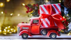 A toy truck with a big present on the back in front of a Christmas tree.